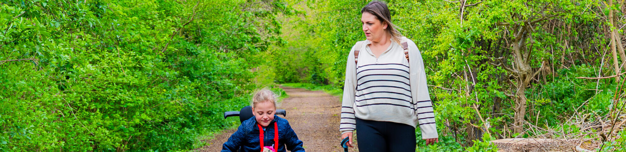 Woman walking beside a girl in a wheel chair. They are in a woodland setting.